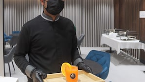 Uma pessoa vestindo uniforme preto carregando uma bandeja com um copo de suco de laranja e uma caixa de madeira.