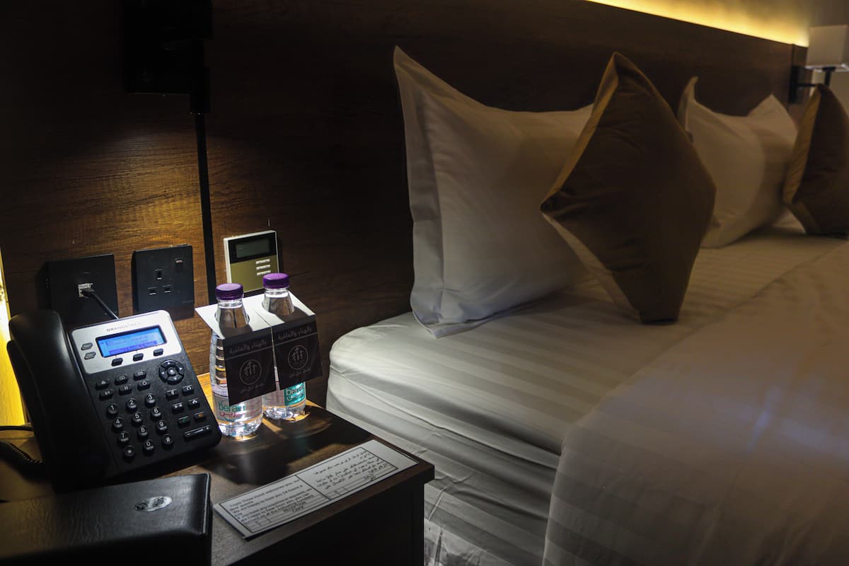 A hotel room with a bed, pillows, a bedside table with a phone, a water bottle, and a digital clock.