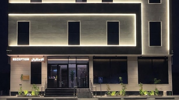 Um hotel moderno com uma placa indicando "TRIPLE THREE HOTEL" e "فندق ثلاثي ثلاثي". Apresenta uma estrutura de vários andares com janelas uniformemente espaçadas e uma área de recepção no nível térreo.