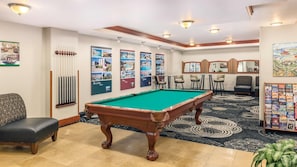 A pool table in a room with a carpeted floor, a pool cue rack, and a seating area with chairs.