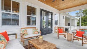 A covered patio area with a wooden ceiling, white walls, and large windows. There are wicker furniture pieces, a wooden table, and a pool visible in the background. - Serene Beach Oasis with Heated Pool (Biloxi)