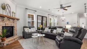 A modern living room with a brick fireplace, leather sofas, a chandelier, and a ceiling fan. - Serene Beach Oasis with Heated Pool (Biloxi)