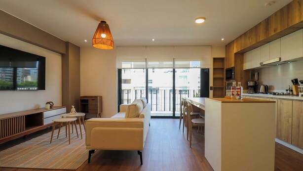 A modern living room with a sofa, coffee table, and TV. It features a kitchen with wooden cabinets and a dining area with chairs. - Engaging 2BR with Cozy Balcony in Miraflores (Miraflores)