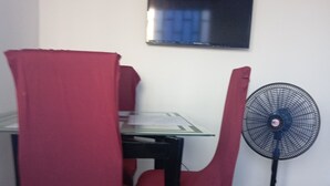 Ein Essbereich mit Tisch und Stühlen, einem an der Wand befestigten Fernseher und einem Standventilator.