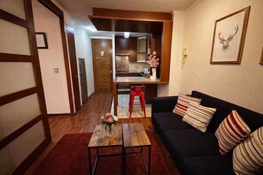 Ein Wohnzimmer mit einer schwarzen Couch, einem Holztisch und einem roten Sessel. Im Hintergrund ist eine Küche mit Kühlschrank und Spüle zu sehen.
