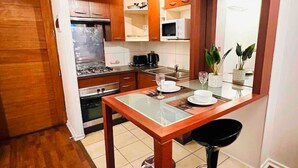 Une cuisine compacte avec des meubles en bois, une cuisinière à gaz, un four à micro-ondes et un coin repas comprenant une table en verre et un tabouret de bar noir.
