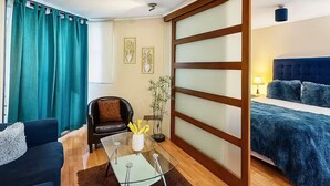 Une chambre d’hôtel moderne comprenant un canapé, une table basse en verre, un fauteuil, un lit avec une tête de lit bleue et une porte coulissante dotée de panneaux en verre dépoli.