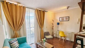 Ein Wohnzimmer mit einem petrolfarbenen Sofa, einem Glastisch, einem gelben Sessel und einem Essbereich mit Tisch und Stühlen.