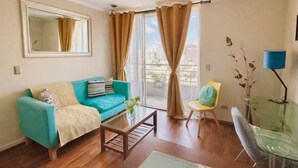 Ein Wohnzimmer mit einem petrolfarbenen Sofa, einem Couchtisch, einem Glastisch mit Lampe, einem Sessel und einem großen Fenster mit Vorhängen.