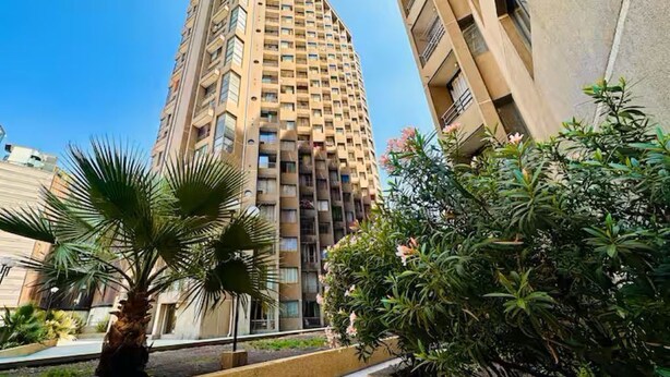Ein hohes, modernes Apartmenthaus mit einem markanten, eckigen Design, umgeben von gepflegter Bepflanzung und blauem Himmel.