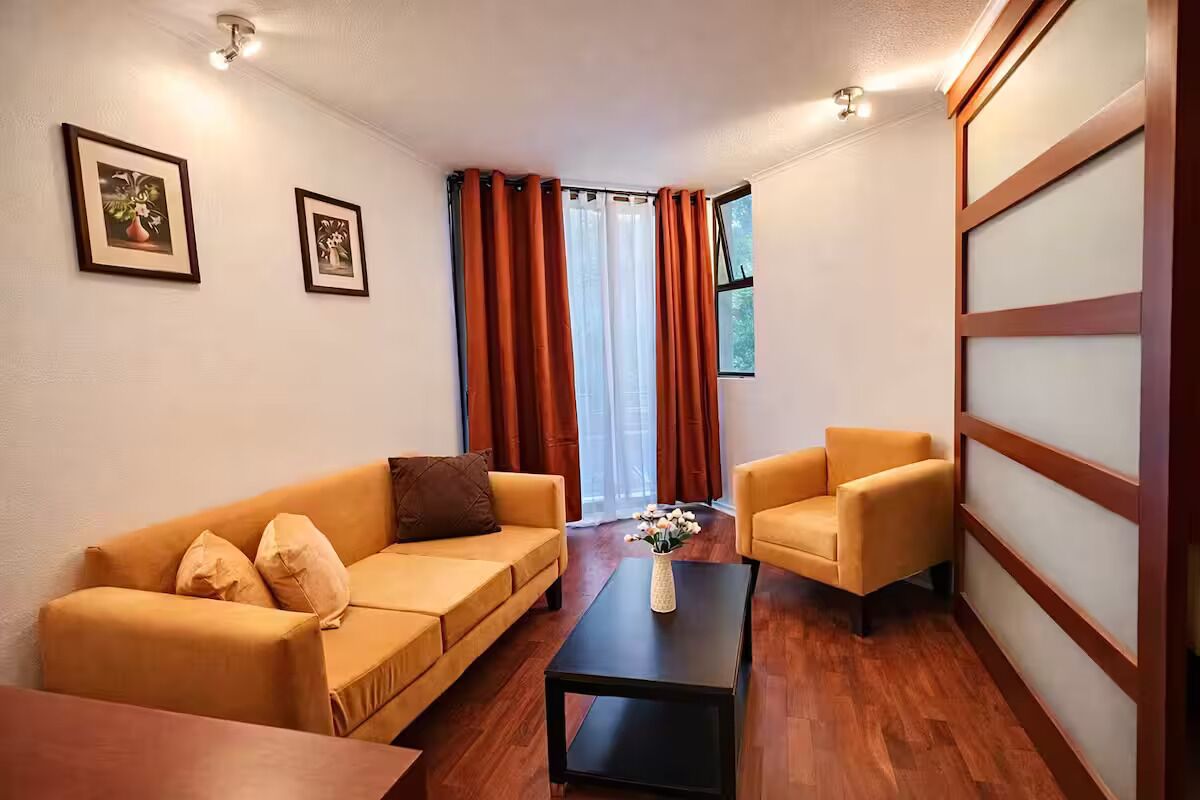 Ein Wohnzimmer mit Sofa, Sessel und Couchtisch. Vorhanden sind Vorhänge, ein Fenster und gerahmte Bilder an der Wand.