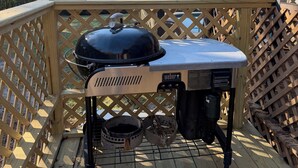 Un barbecue Weber sur une terrasse en bois dotée d’une rambarde en treillis, une cheminée, et une vue sur des arbres et une maison en arrière-plan.