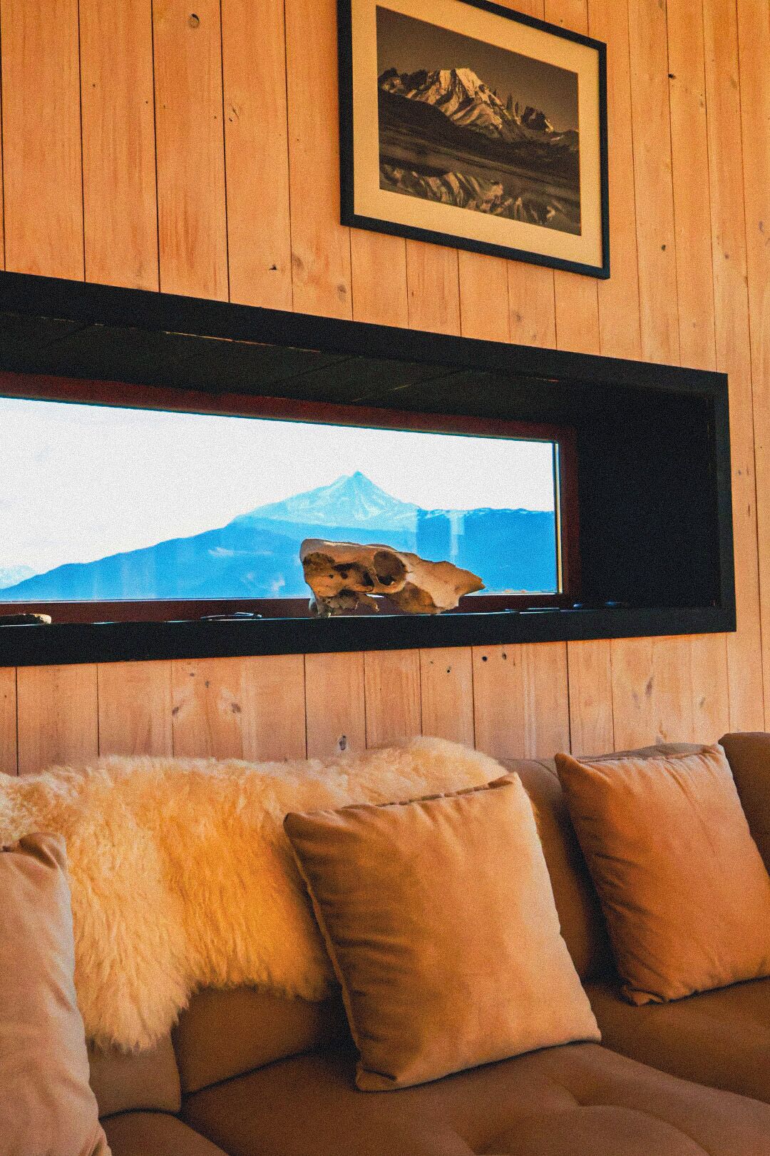 A wooden interior with a sofa, pillows, a framed picture of mountains, and a window with a mountain view.