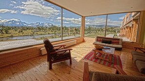 Una habitación amplia con suelo de madera, un gran ventanal con vistas a la montaña, una zona de estar acogedora con sofá y sillones, y una estantería de madera.