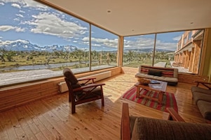 Una habitación amplia con suelo de madera, un gran ventanal con vistas a la montaña, una zona de estar acogedora con sofá y sillones, y una estantería de madera.