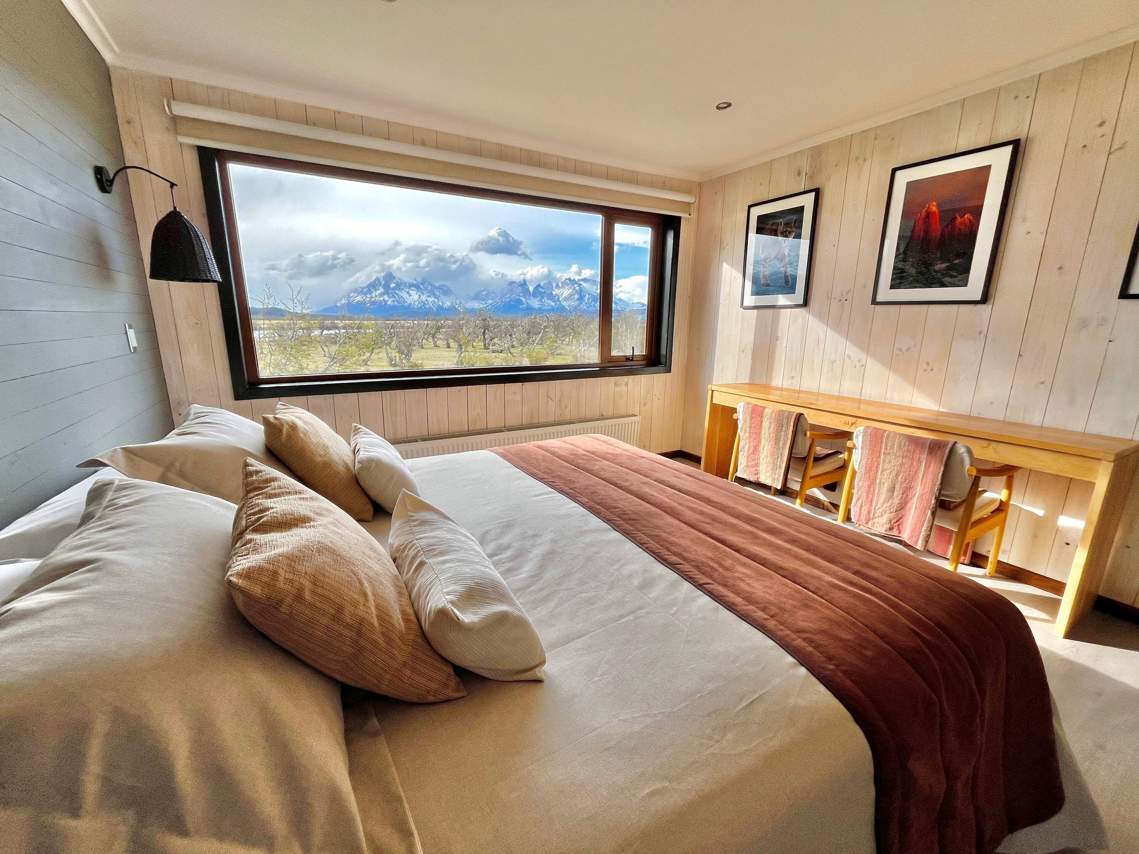 A bedroom with a large window, a bed with pillows, a bedside table, and a chair.