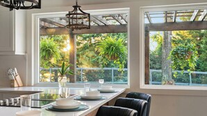 Cocina con una isla central grande, encimeras blancas y sillas negras. Dos ventanas ofrecen vistas a zonas verdes y una pérgola de madera.