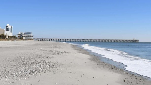 A beach with a long pier extending into the ocean, a building with a white facade, and a clear blue sky. - NEW, Large 2BR in Ocean Greens,Great Location,Pool (North Myrtle Beach)