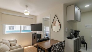 Un espacio habitable compacto con sofá, mesa de comedor, televisor, zona de cocina con cafetera y ventilador de techo.