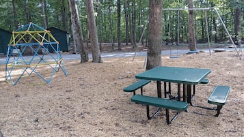 Un parque infantil con una estructura para escalar y mesas de picnic en un área boscosa.