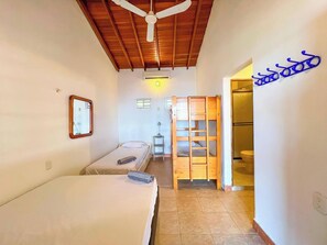 A small, well-lit room with two single beds, a ceiling fan, a wooden wardrobe, and a bathroom with a toilet and sink.