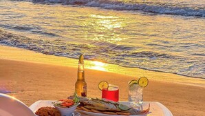 Ein Tisch am Strand mit Speisen, Getränken und einer Flasche, vor einer Sonnenuntergangsansicht.