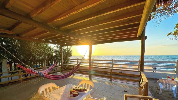Eine Terrasse direkt am Strand mit Tisch für zwei Personen, einer Hängematte und Blick auf das Meer bei Sonnenuntergang.