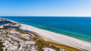 Un paysage côtier avec une plage de sable, des eaux d’un bleu limpide et une ligne de côte lointaine où se dressent des bâtiments.