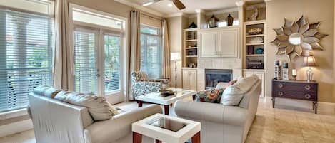 A living room with two sofas, a coffee table, and a fireplace. There are large windows with blinds, a ceiling fan, and a decorative wall piece.