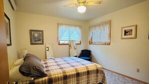 A bedroom with a bed, a ceiling fan, two windows with curtains, and a carpeted floor.