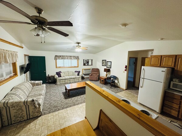 A living room with a sofa, armchair, and coffee table. There is a ceiling fan, a window with blinds, and a door.