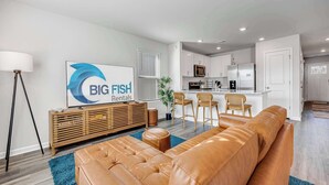Ein modernes Wohnzimmer mit einer Ledersofa, einem Fernseher, der 'BIG FISH Rentals' anzeigt, einer Holztruhe und einer Küchenzeile mit Mikrowelle und Kühlschrank.