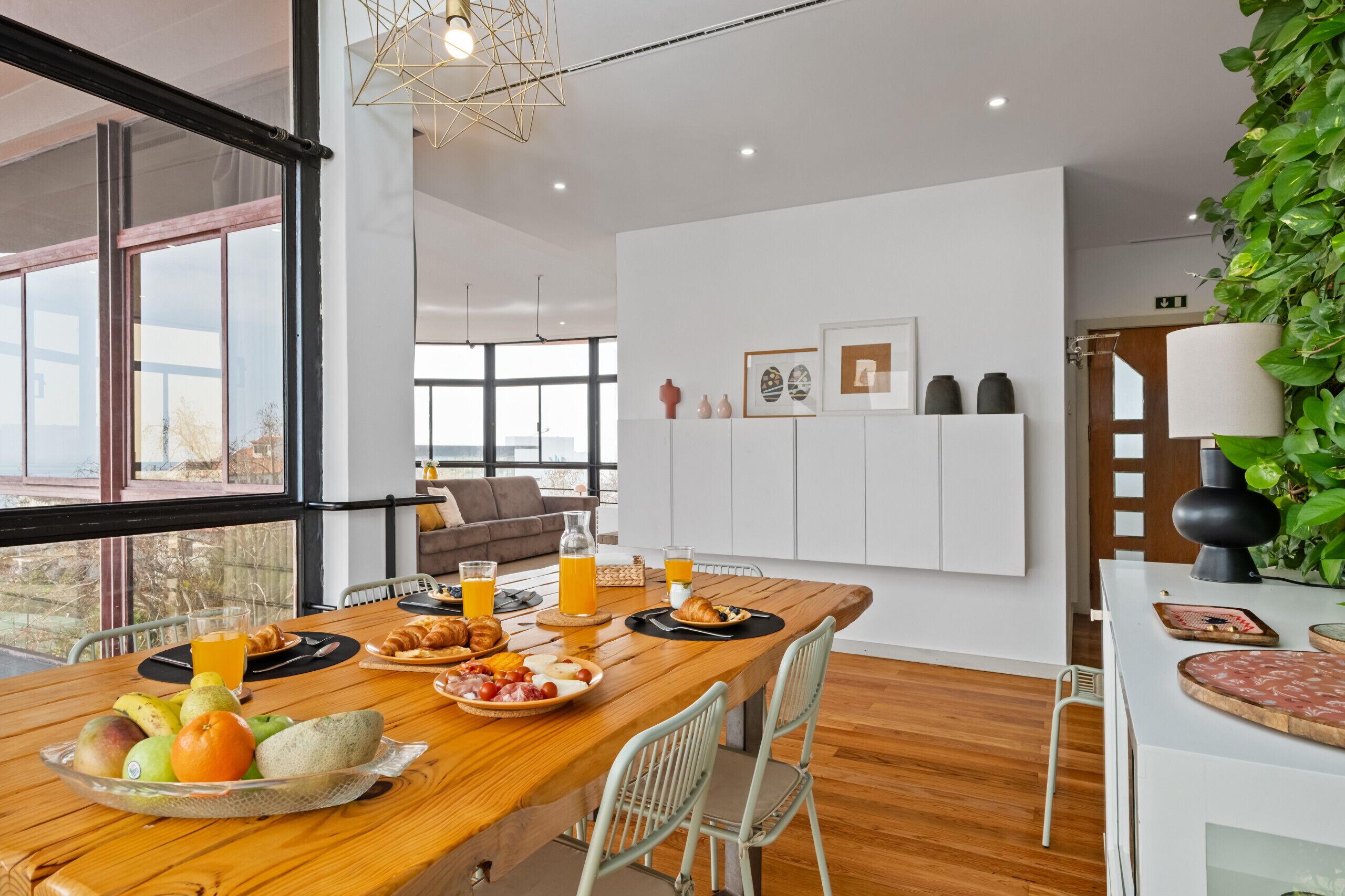 A modern dining area with a wooden table set for breakfast, large windows, and a view of the outdoors.