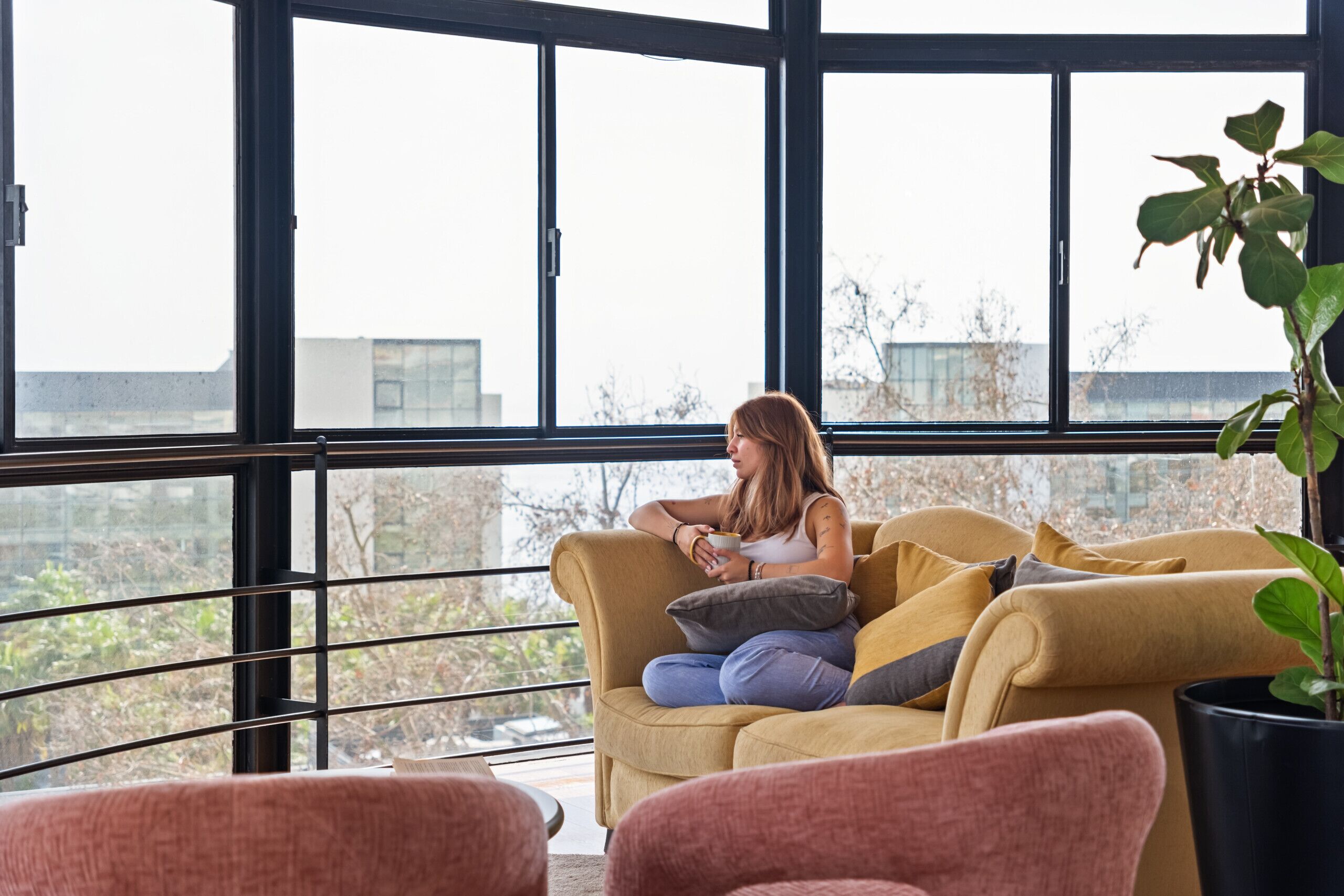 A woman sitting on a yellow sofa with a view of a cityscape through large windows.