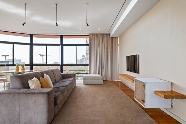 Un salon moderne avec un canapé gris, une grande fenêtre, un téléviseur fixé au mur et une étagère en bois.