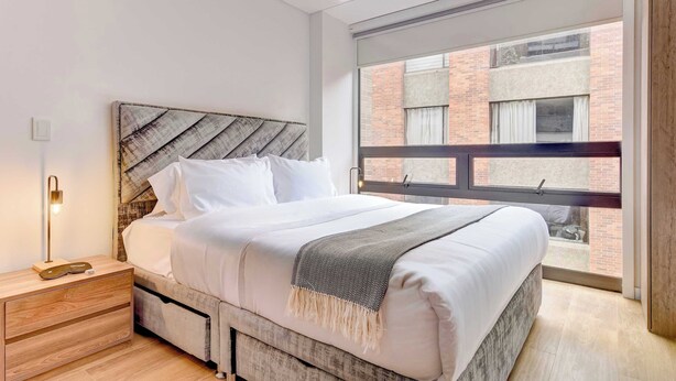 Ein modernes Schlafzimmer mit einem großen Bett, einem Nachttisch aus Holz, einer Nachttischlampe und Blick auf ein Backsteingebäude durch ein Fenster.