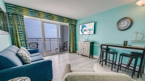 Un salon moderne avec un canapé, des fauteuils, une table basse et un coin repas avec des tabourets. Une grande fenêtre offre une vue sur le balcon, et une enseigne indiquant « Big Fish Rentals » est visible sur le mur.
