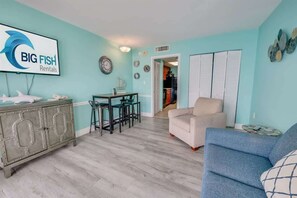 A living room with teal walls, a dining area with a table and chairs, a sofa, and a cabinet with a shark decoration. - 1BR High Floor, Great Views, Camelot Resort, Pools (Myrtle Beach)