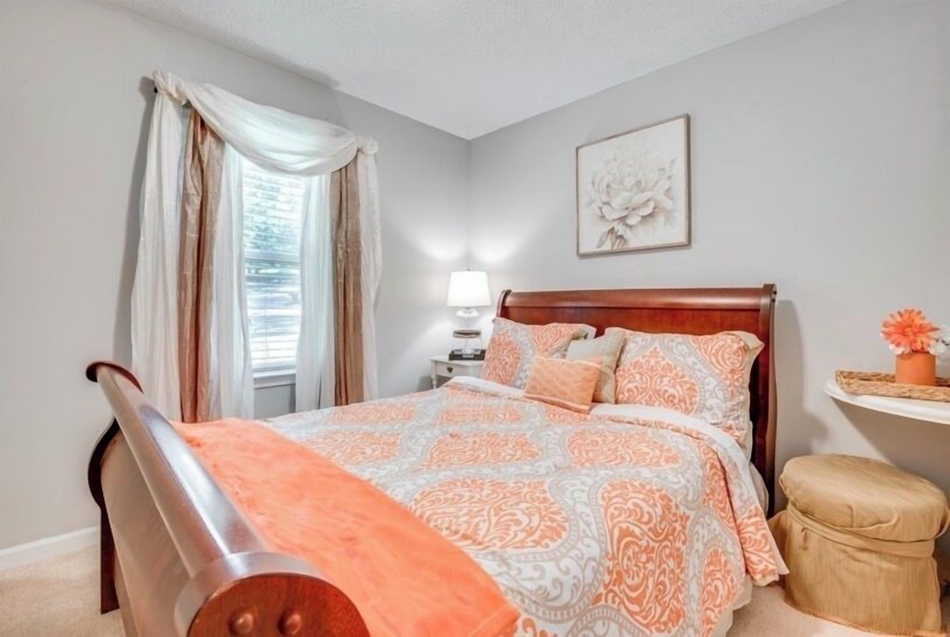 Un dormitorio con cama, mesita de noche, silla, una pequeña mesa y ventana con cortinas.