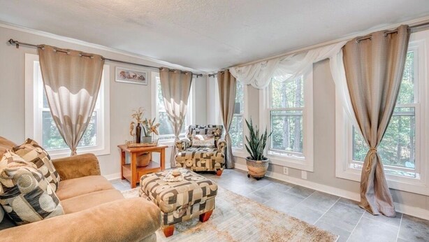 A spacious living room with a beige sofa, a patterned ottoman, and a wooden side table. There are two windows with beige curtains, a potted plant, and a framed picture on the wall. - 3-bedroom house in awesome Ellenwood with cool AC breeze (Ellenwood)
