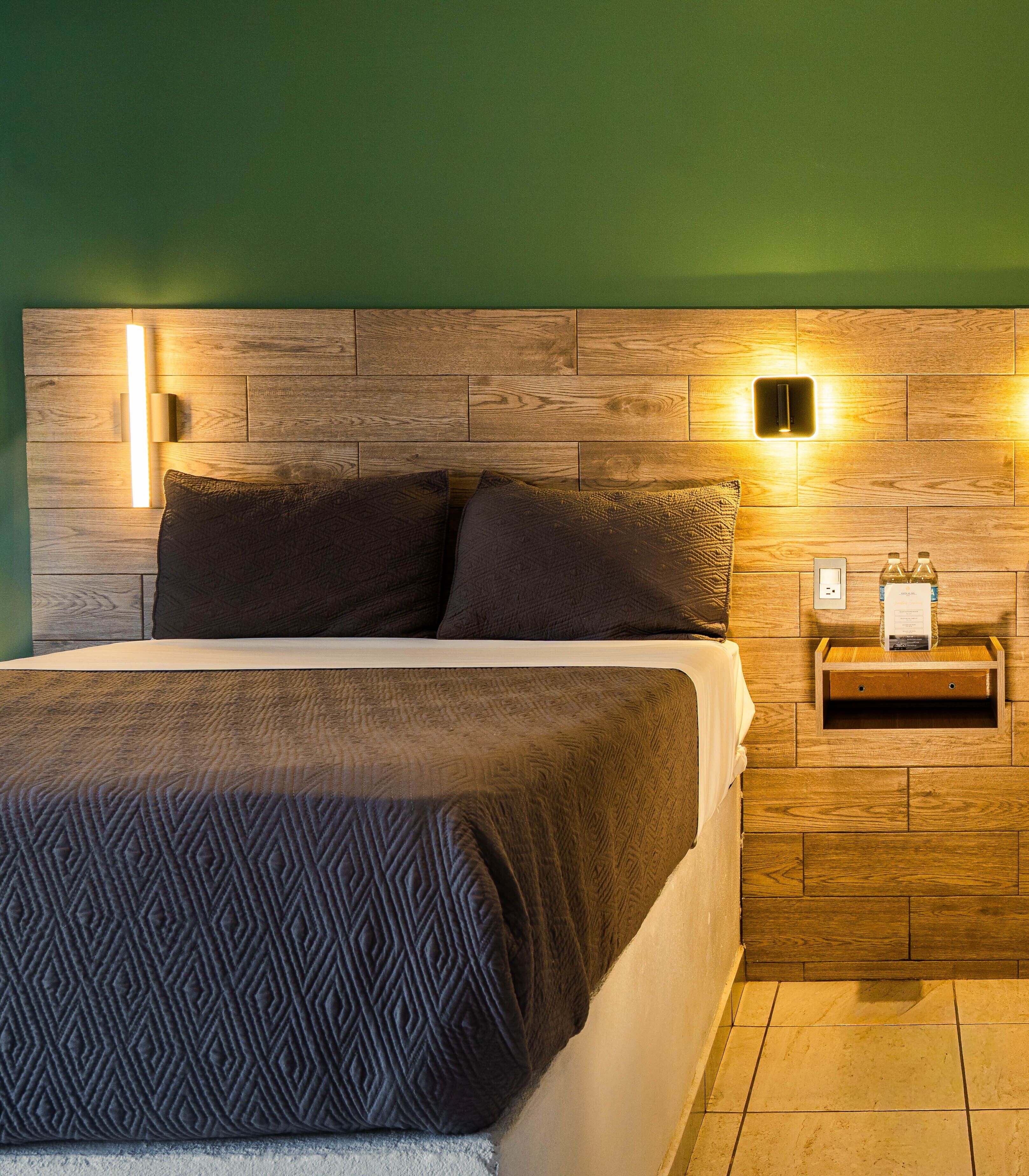 Una cama con una colcha estampada, un cabecero de madera con luces fijadas a la pared y una mesita de noche con una botella y una caja.