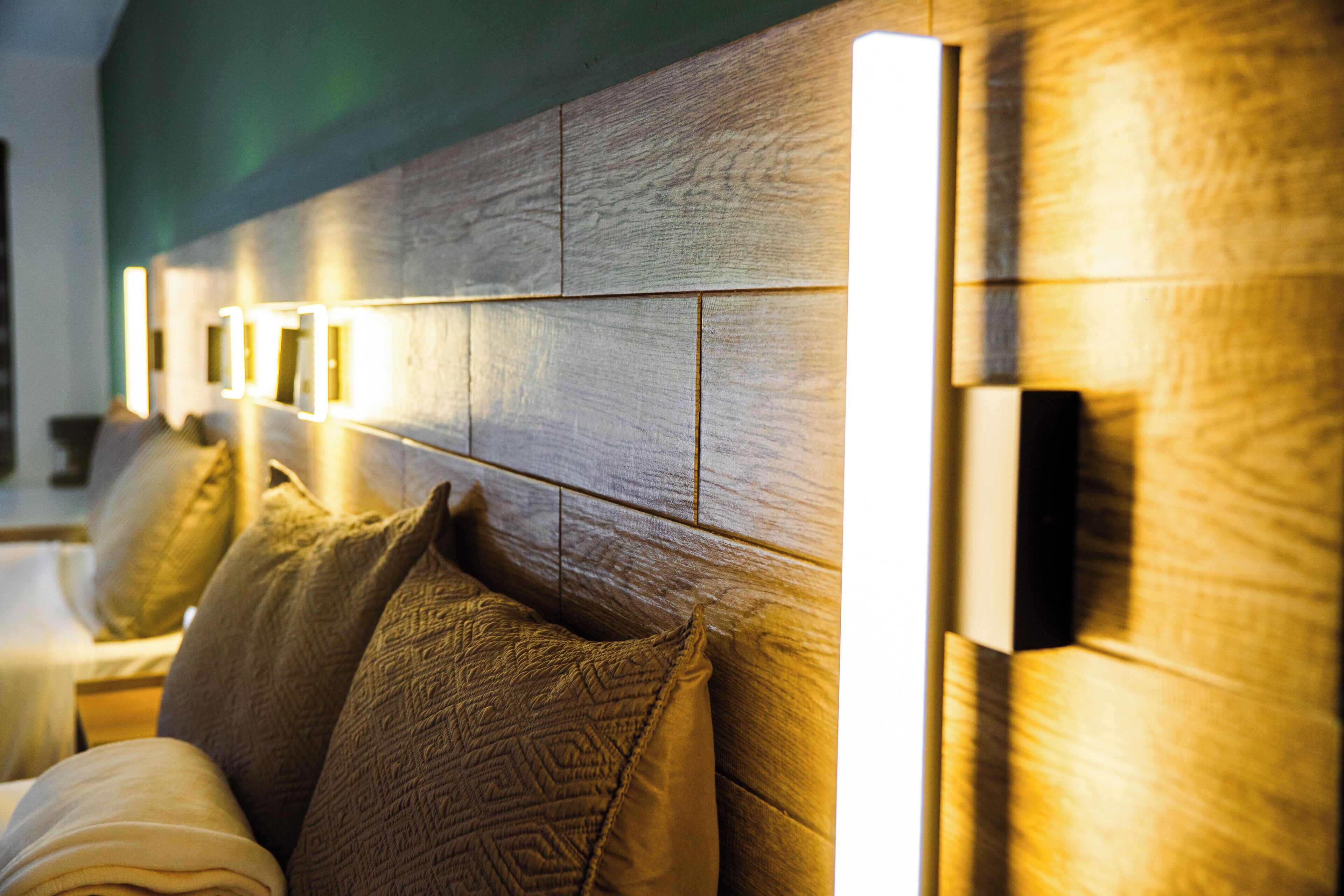 Una camera d'albergo con testiera in legno, numerosi cuscini e illuminazione a parete.