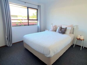 A bedroom with a large bed, a nightstand, a window with curtains, and a view of the outside. - Serene Family Holiday Home, Pillans Point - Tga (Tauranga)