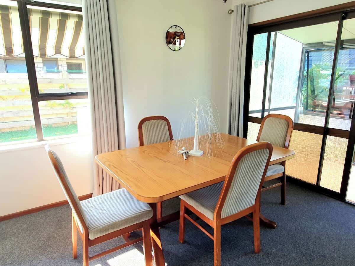 Una habitación con una mesa de madera y cuatro sillas, un reloj en la pared y ventanas grandes.