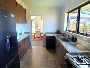 A compact kitchen with a refrigerator, stove, and sink, leading to a dining area with a chair and table. - Serene Family Holiday Home, Pillans Point - Tga (Tauranga)
