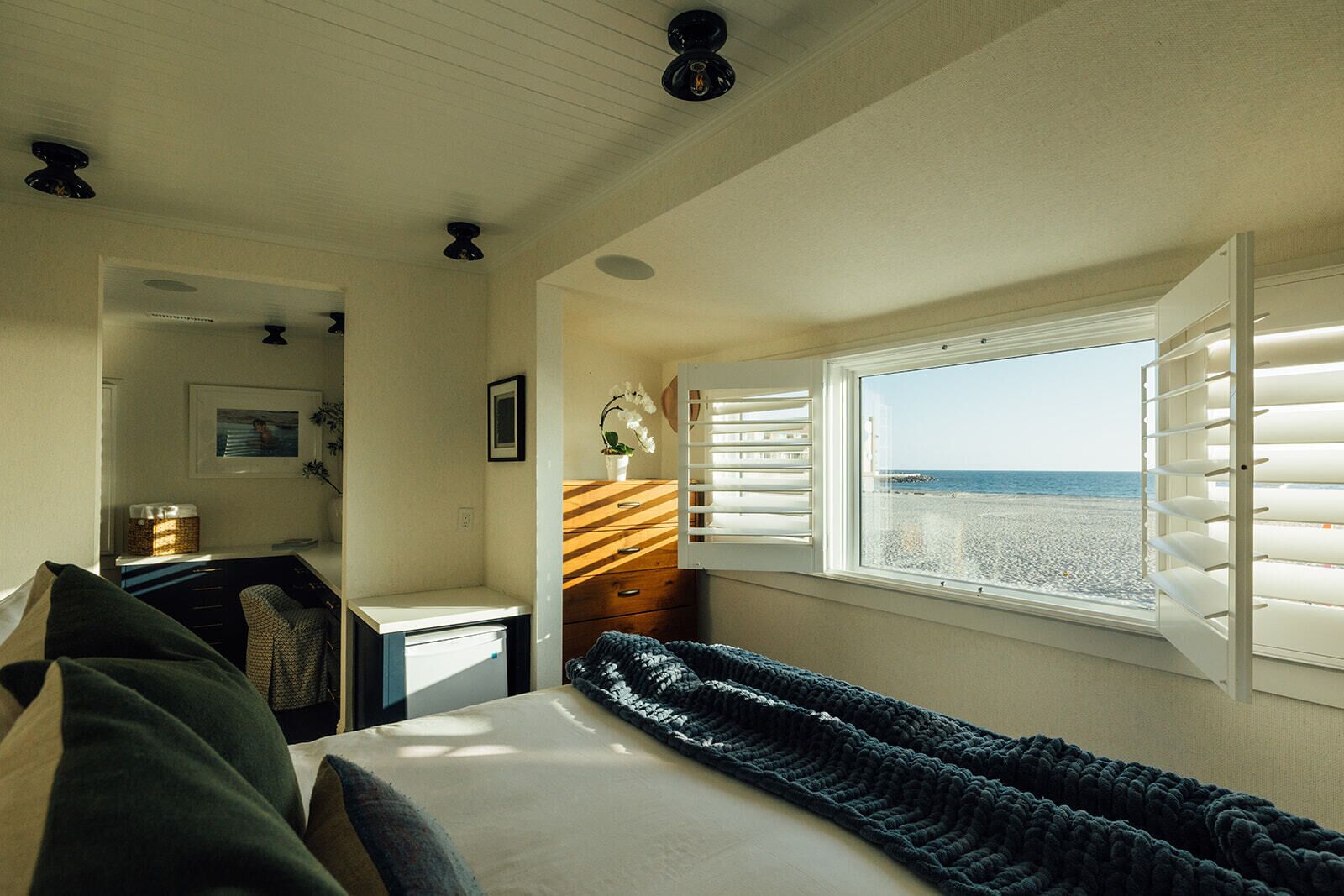 Et soveværelse med seng, kommode, vindue med udsigt til stranden og ventilator i loftet.