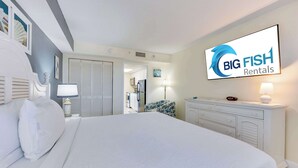A hotel room with a large bed, a dresser, a TV displaying 'Big Fish Rentals', and a view into the kitchen area. - Honeymoon Suite with Direct Oceanfront View,Pools! (Myrtle Beach)