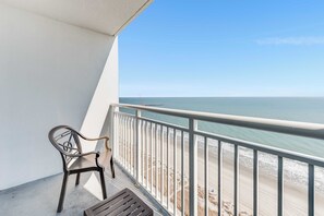 Un balcon avec une chaise en métal et une table en bois, donnant sur une plage sous un ciel dégagé.