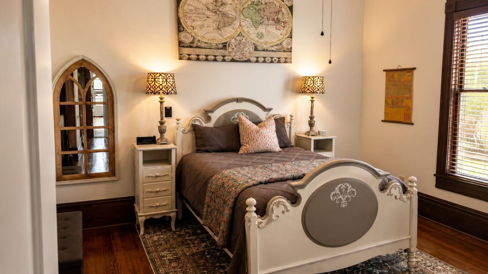 Een slaapkamer met een groot bed, twee nachtkastjes met lampen, een decoratieve kaart aan de muur, en een raam met jaloezieën.