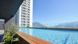 Ein Pool auf dem Dach mit klarem, blauem Wasser, umgeben von einer Holzterrasse und Pflanzen. Im Hintergrund ein hohes Gebäude und ein Gebirge unter blauem Himmel.